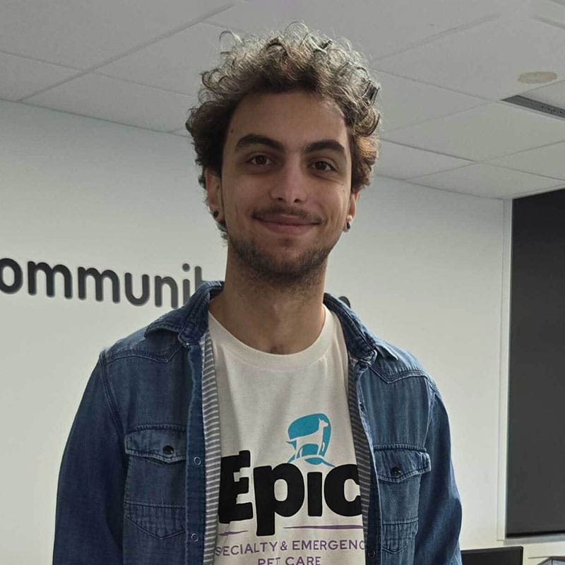 A young man with curly hair is smiling at the camera. He is wearing a denim shirt over a t-shirt that says "Epic Specialty & Emergency Pet Care," highlighting his role at a leading Emergency Vet clinic. The background features a white wall with black text.