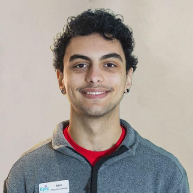 A young person with short, curly black hair, wearing a gray zip-up jacket over a red shirt, smiles at the camera. They have small hoop earrings, a name tag that reads "Adam," and look ready to assist at the Pet ER. The background is plain and light-colored.