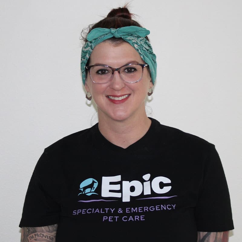 A person with glasses and a green headscarf smiles at the camera. They are wearing a black shirt that says “Epic Specialty & Emergency Pet Care”—perfect for an emergency vet or dedicated veterinarian.