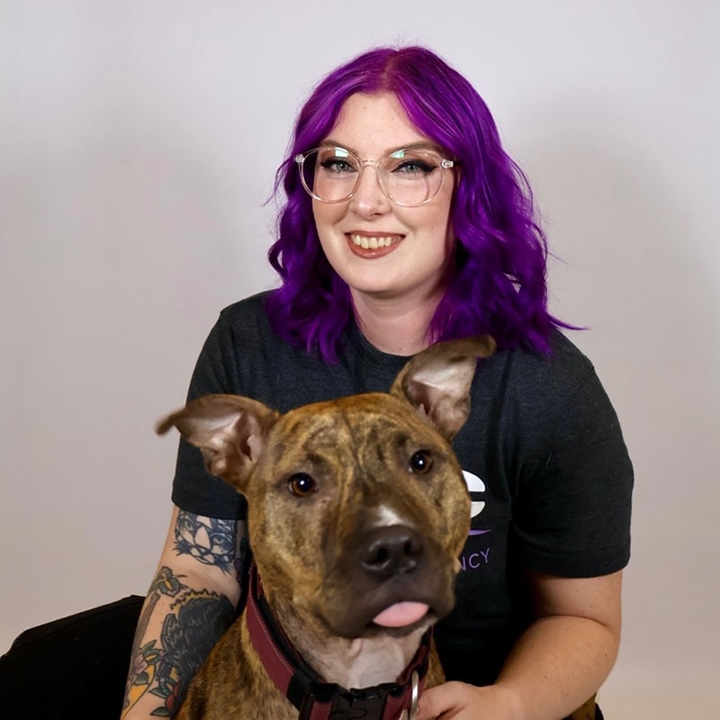 A smiling person with purple hair and glasses sits in front of a plain background, holding a brown and black dog with pointy ears and a pink tongue sticking out.