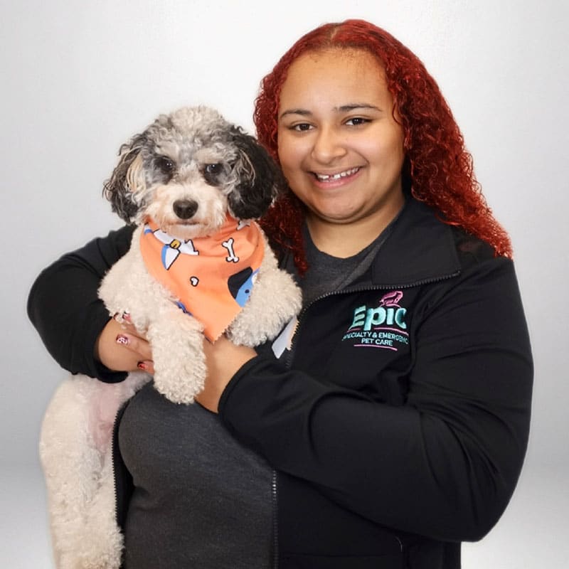 Smiling person with curly red hair holds a black, white, and gray fluffy dog wearing an orange bandana with dog-themed designs. The person wears a black jacket with an "Epic Pet Care" logo on it, against a plain background.