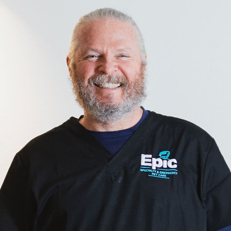 A smiling bearded man with gray hair, wearing a black "Epic Specialty & Emergency Pet Care" shirt, stands against a plain white background—ready to help as your trusted emergency vet.