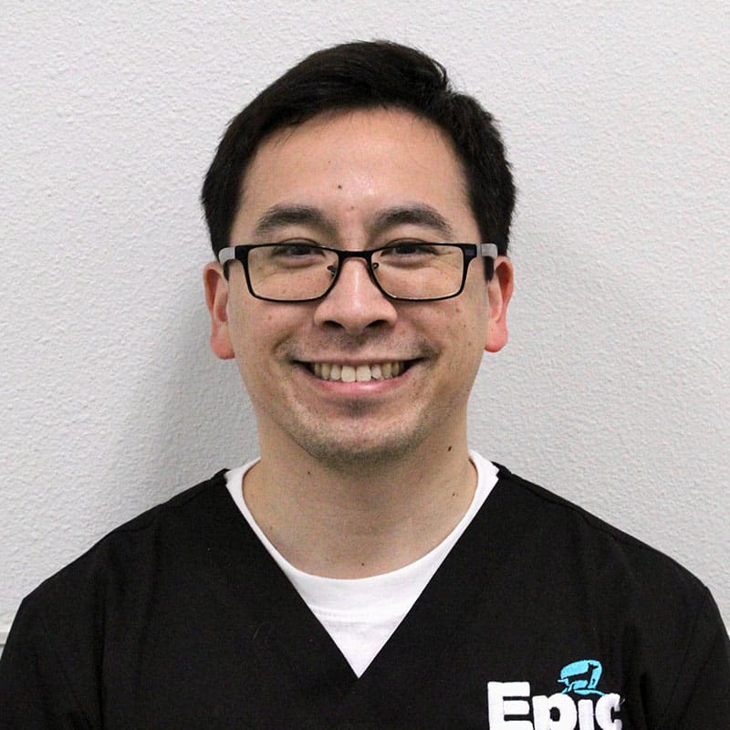 A man with short dark hair and glasses smiles at the camera while wearing black scrubs with "Epic" written on the chest, standing in front of a plain light-colored wall—ready to help as an experienced Emergency Vet.