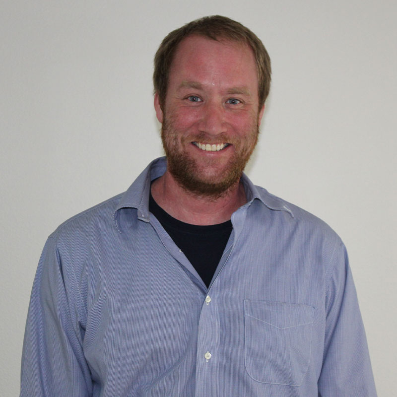 A man with short brown hair and a beard smiles at the camera. He is wearing a light blue button-up shirt over a black t-shirt, standing in front of a plain white background, ready to help as your trusted Pet ER veterinarian.
