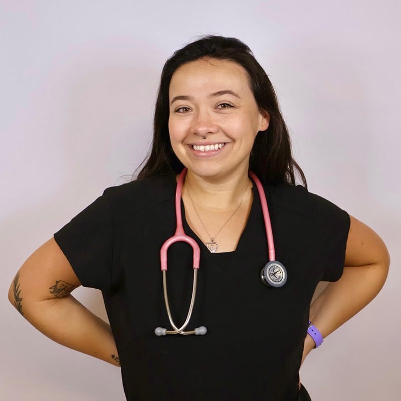 A woman in black scrubs smiles at the camera with her hands on her hips. She has a pink stethoscope around her neck and wears a purple watch. The background is plain and light-colored.