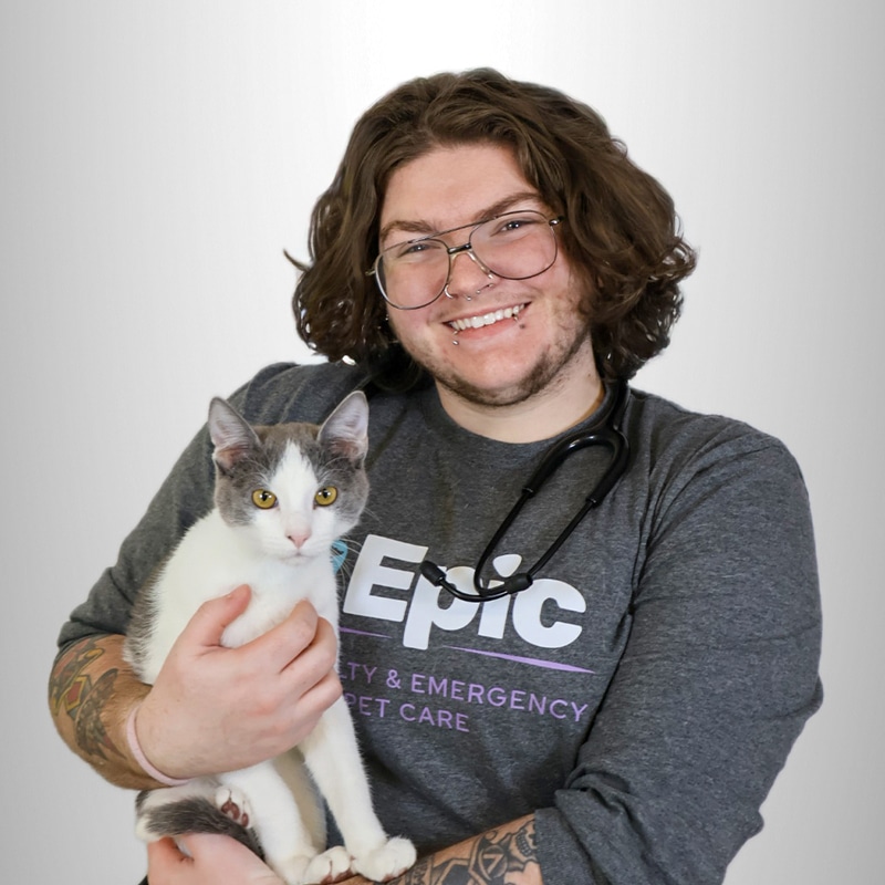 Smiling person with long curly hair and glasses, wearing a gray “Epic Specialty & Emergency Pet Care” shirt, holds a white and gray cat while a stethoscope hangs around their neck.