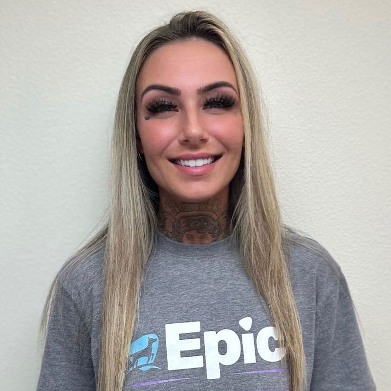 A woman with long blonde hair and visible neck tattoos smiles at the camera, wearing a gray "Epic" t-shirt. She stands in front of a plain light-colored wall, looking confident—like she could handle any Emergency Vet situation.