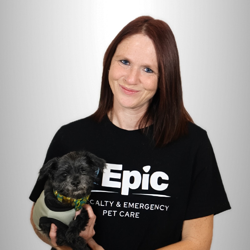 Smiling woman with straight, dark red hair holds a small black dog. She wears a black “Epic Specialty & Emergency Pet Care” t-shirt. The background is plain light gray.