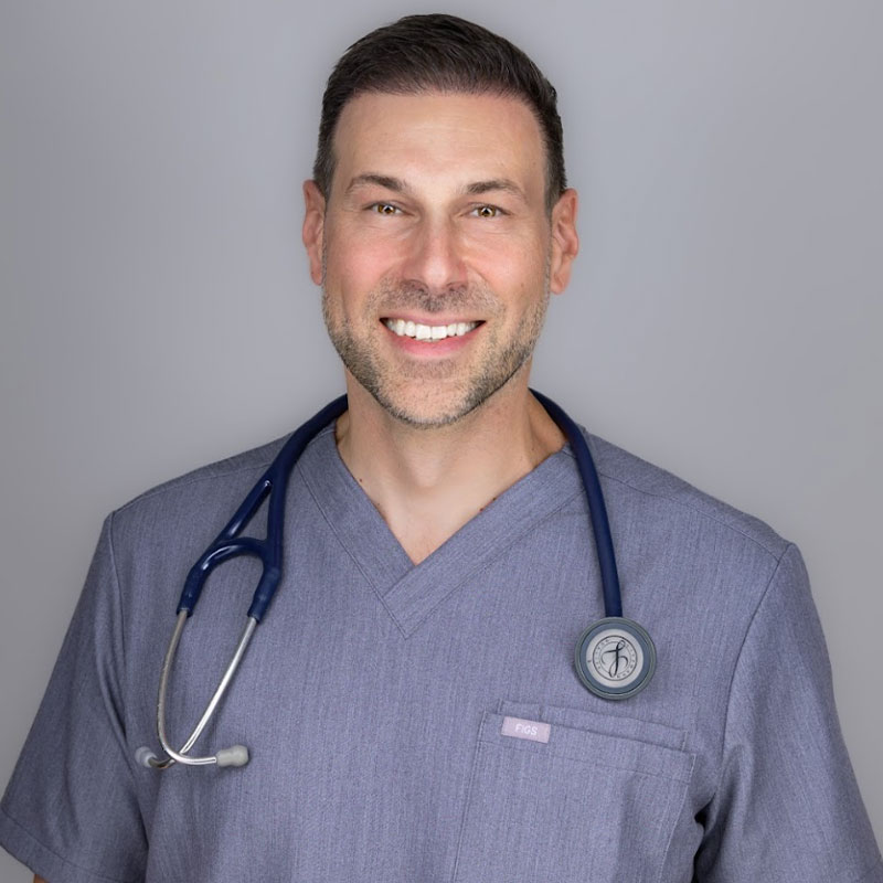 A smiling veterinarian wearing gray scrubs and a stethoscope around his neck stands against a plain gray background.