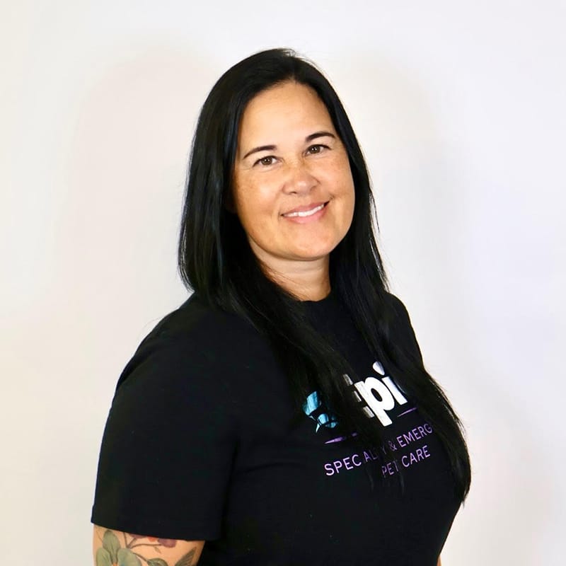 A woman with long dark hair smiles at the camera, wearing a black t-shirt with text on it, standing against a plain white background.