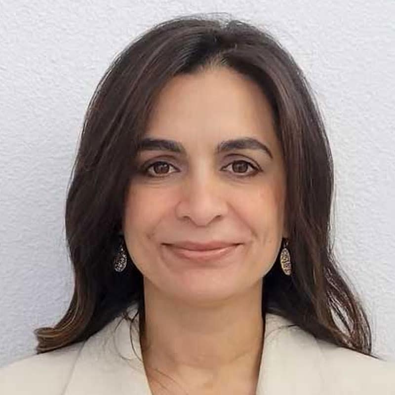 A woman with long dark hair, wearing a light-colored blazer and dangling earrings, smiles softly in front of a plain white background, ready to assist as your trusted emergency vet.