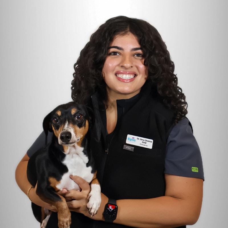 A smiling woman with curly hair holds a black and brown dog. She wears a uniform and a name badge, standing against a plain white background.