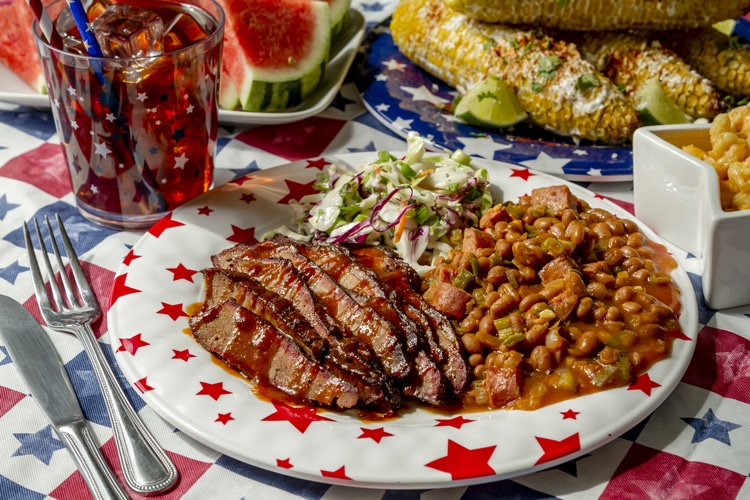 A patriotic plate with BBQ brisket in sauce, baked beans, and coleslaw on a star-patterned dish, set on a red, white, and blue tablecloth. Sides include corn on the cob, watermelon slices, iced drink, and macaroni and cheese.