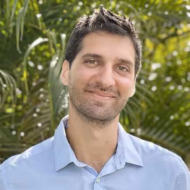 A man with short dark hair and a trimmed beard smiles gently at the camera, wearing a light blue collared shirt. Green foliage is blurred in the background, suggesting an outdoor setting, possibly near a Vet or Pet ER clinic.