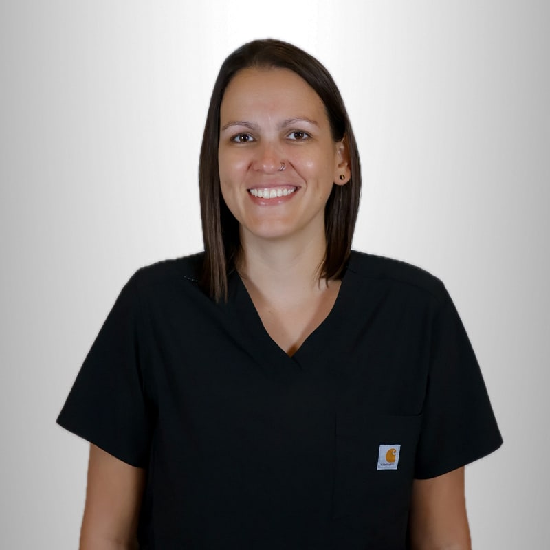 A person with straight brown hair, wearing black medical scrubs, stands smiling in front of a plain light gray background.