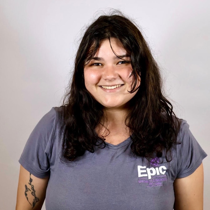 A person with long dark hair smiles at the camera, wearing a gray “Epic Special Education” T-shirt. They have a visible arm tattoo and are standing against a plain light background.