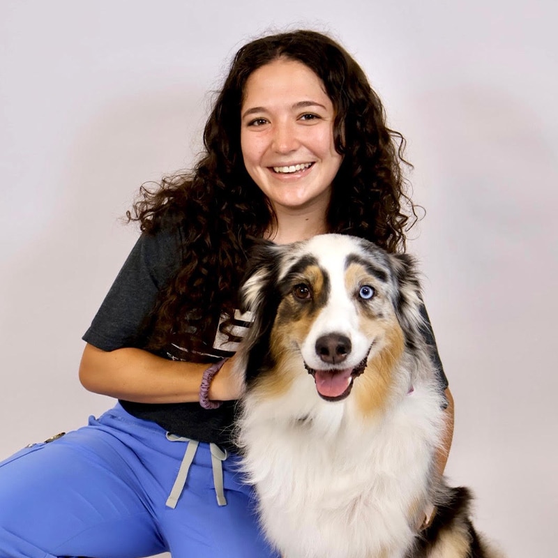 A smiling person with curly hair, wearing a dark t-shirt and blue pants, poses next to a fluffy dog with one brown eye and one blue eye against a plain light background.